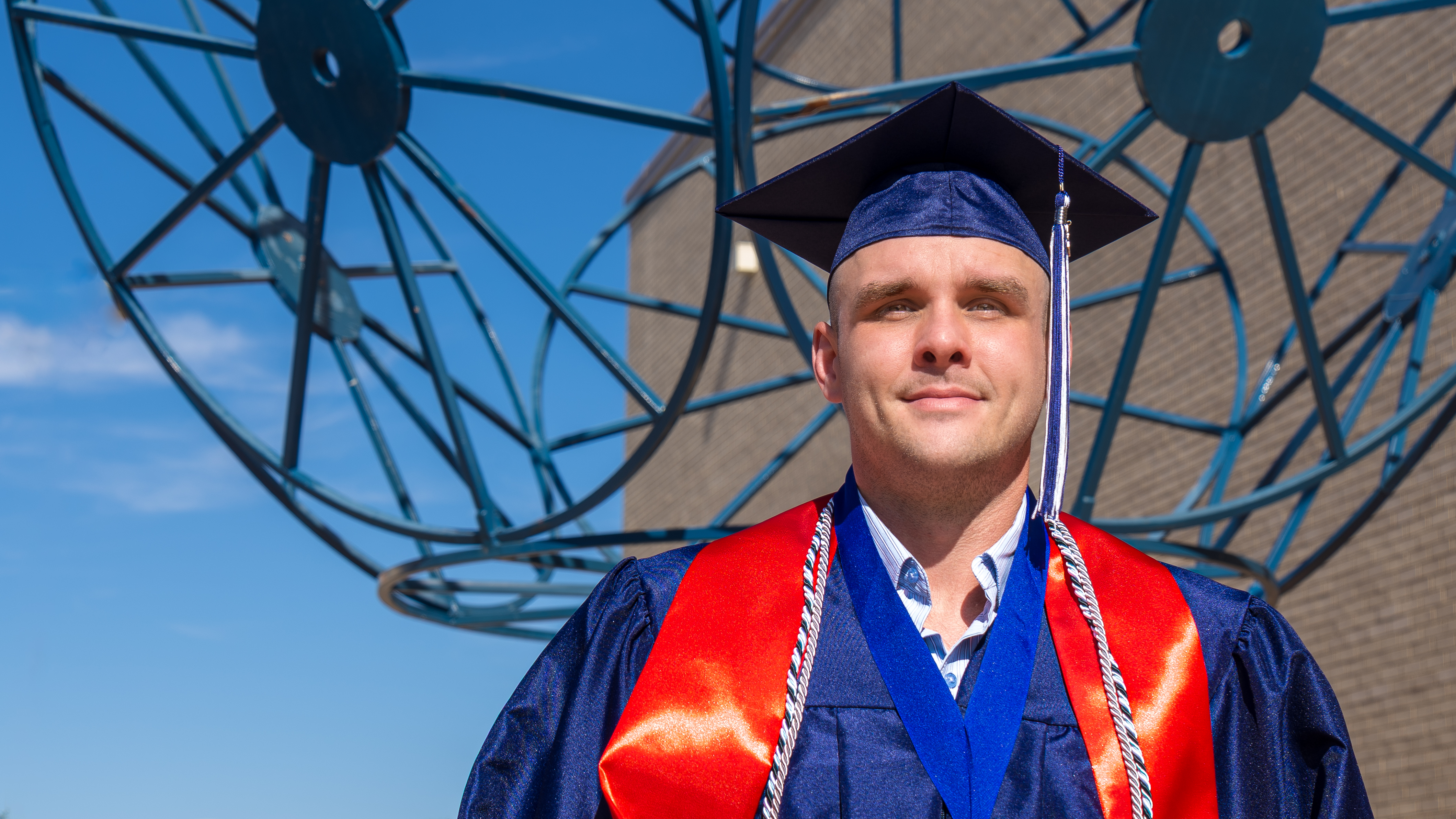 A smiling man in a navy graduation cap and gown with a red stole and silver honors cords, standing in front of a large blue metal sculpture.