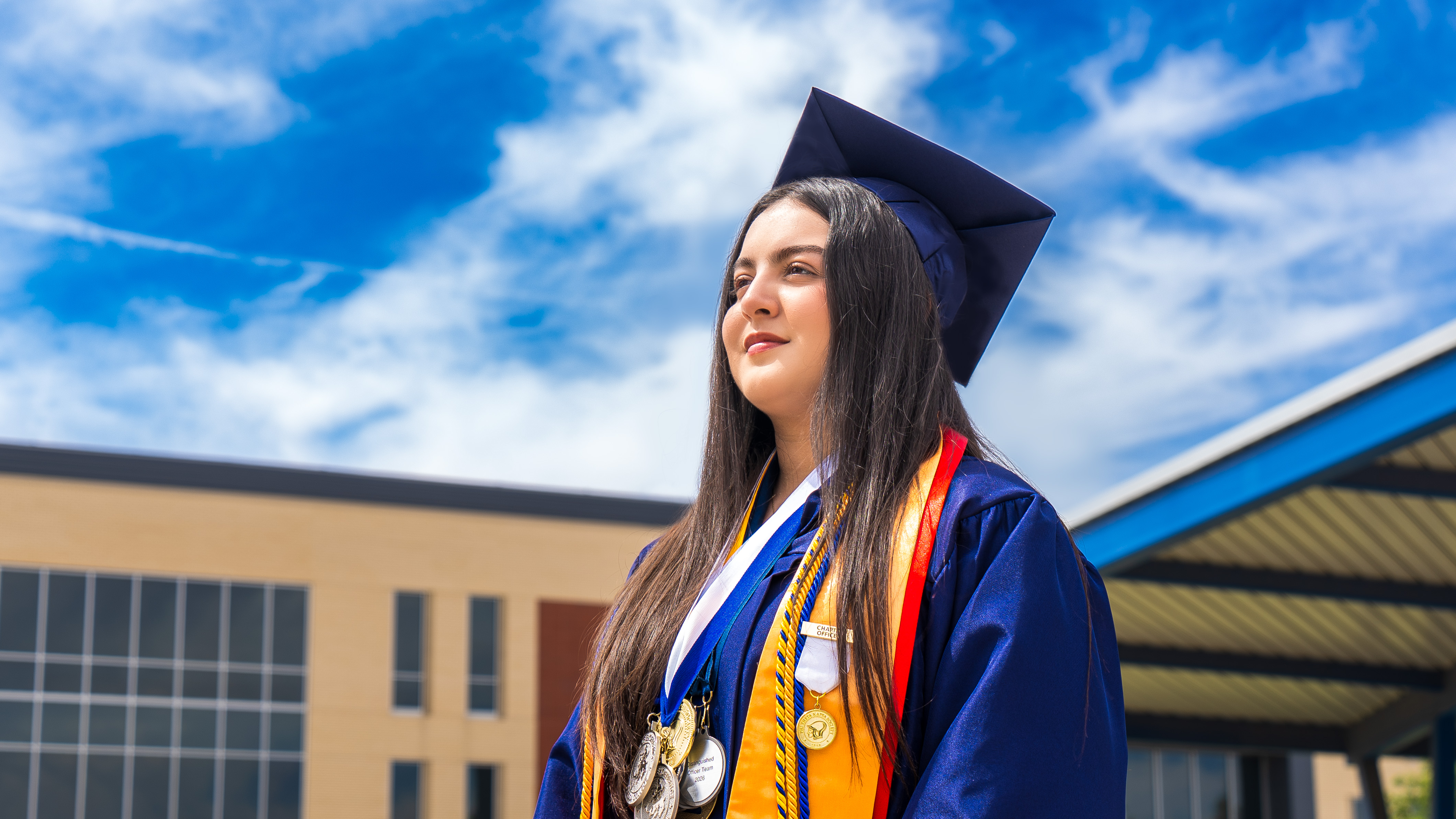A young woman in a blue graduation cap and gown stands outside a school building, wearing multiple academic medals and a gold stole against a bright, cloudy sky.